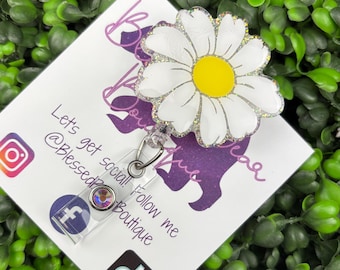 Puede incluir: Un portagafete retráctil plateado con una margarita blanca con purpurina y un centro amarillo. El portagafete tiene un rectángulo de plástico transparente con una pequeña gema iridiscente y la letra "F". El portagafete está unido a una tarjeta blanca con el texto "Let's get social! Follow me @BlessedzBoutique" e iconos de redes sociales para Instagram, Facebook y TikTok.
