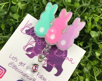 May include: A retractable badge reel featuring three colorful bunny designs in mint green, pink, and lavender. Each bunny has a white tail and is outlined with glitter. The reel has a pink gem and a clear plastic strap.