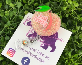 May include: A pink glitter peach with a green leaf and the text "Just Peachy" on it. The peach is attached to a clear badge holder with a silver clip. The badge holder is on a white card with the text "Blessed Bear Boutique" and social media icons.