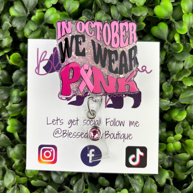 A flat lay image displays a badge reel with the words "IN OCTOBER WE WEAR PINK" in bold, black and pink lettering. A pink ribbon symbol is incorporated into the design. The background of the badge reel is a light pink glitter.