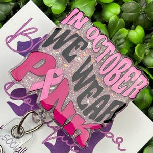 A close-up shot of a hand holding a rectangular retractable badge reel against a background of green artificial leaves. The badge reel has a light pink glitter background. The text "IN OCTOBER WE WEAR PINK" is written in black and bold pink font with a large pink awareness ribbon.