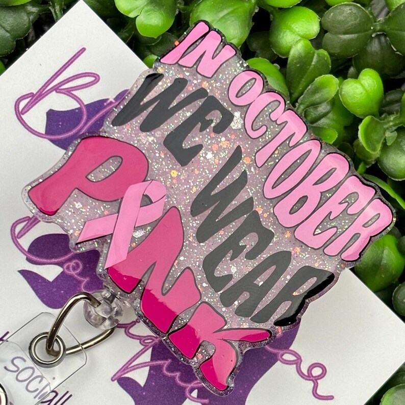A close-up shot of a hand holding a rectangular retractable badge reel against a background of green artificial leaves. The badge reel has a light pink glitter background. The text "IN OCTOBER WE WEAR PINK" is written in black and bold pink font with a large pink awareness ribbon.