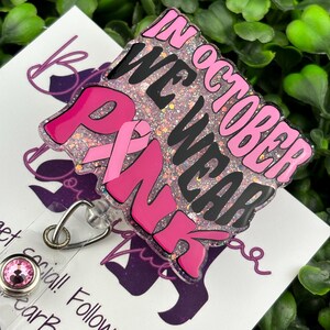 A flat lay image displays a badge reel with the words "IN OCTOBER WE WEAR PINK" in bold, black and pink lettering. A pink ribbon symbol is incorporated into the design. The background of the badge reel is a light pink glitter.