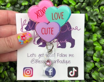 Op de afbeelding: Een badge reel met drie hartvormige bedels in roze, teal en paars, elk met een woord: "XOXO", "LOVE" en "CUTE". De reel heeft een doorzichtige plastic houder en een roze edelsteen. De achtergrond is een groene bladertextuur.