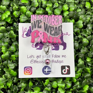 A close-up shot of a hand holding a rectangular retractable badge reel against a background of green artificial leaves. The badge reel has a light pink glitter background. The text "IN OCTOBER WE WEAR PINK" is written in black and bold pink font with a large pink awareness ribbon.