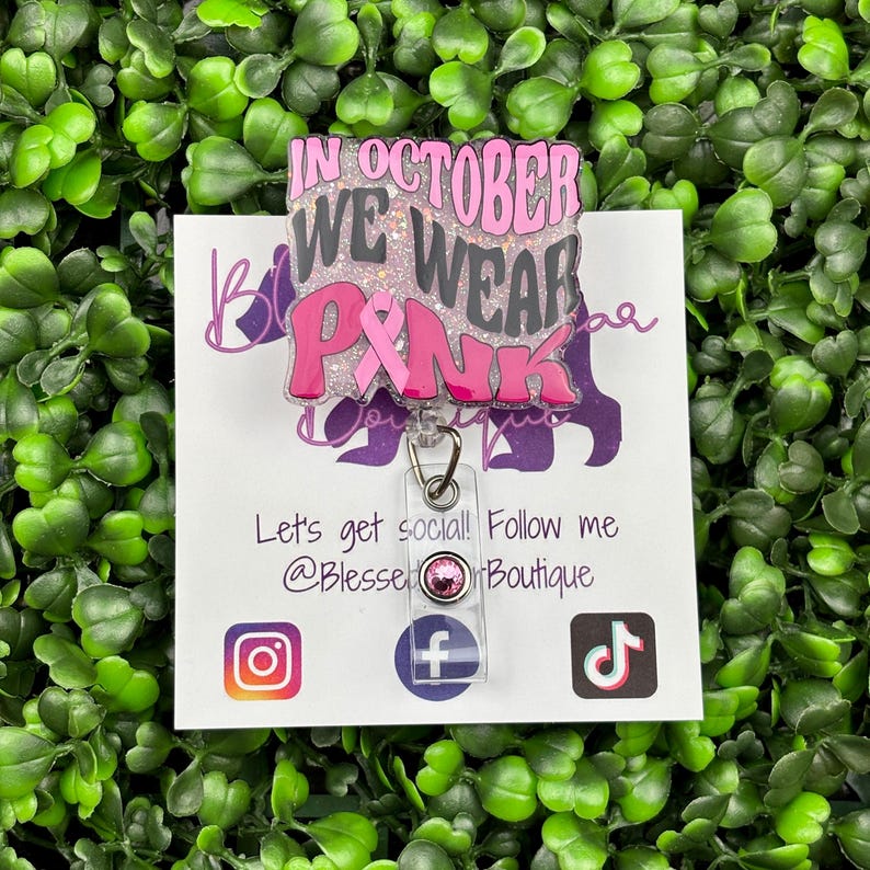 A close-up shot of a hand holding a rectangular retractable badge reel against a background of green artificial leaves. The badge reel has a light pink glitter background. The text "IN OCTOBER WE WEAR PINK" is written in black and bold pink font with a large pink awareness ribbon.