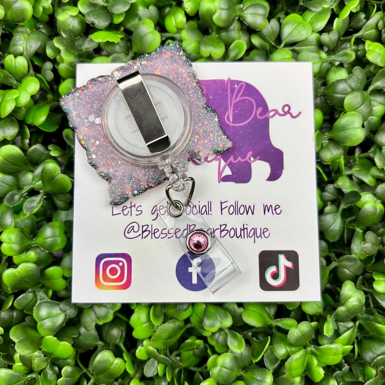 A close-up shot of a hand holding a rectangular retractable badge reel against a background of green artificial leaves. The badge reel has a light pink glitter background. The text "IN OCTOBER WE WEAR PINK" is written in black and bold pink font with a large pink awareness ribbon.