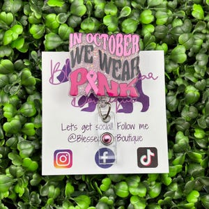 A close-up shot of a hand holding a rectangular retractable badge reel against a background of green artificial leaves. The badge reel has a light pink glitter background. The text "IN OCTOBER WE WEAR PINK" is written in black and bold pink font with a large pink awareness ribbon.