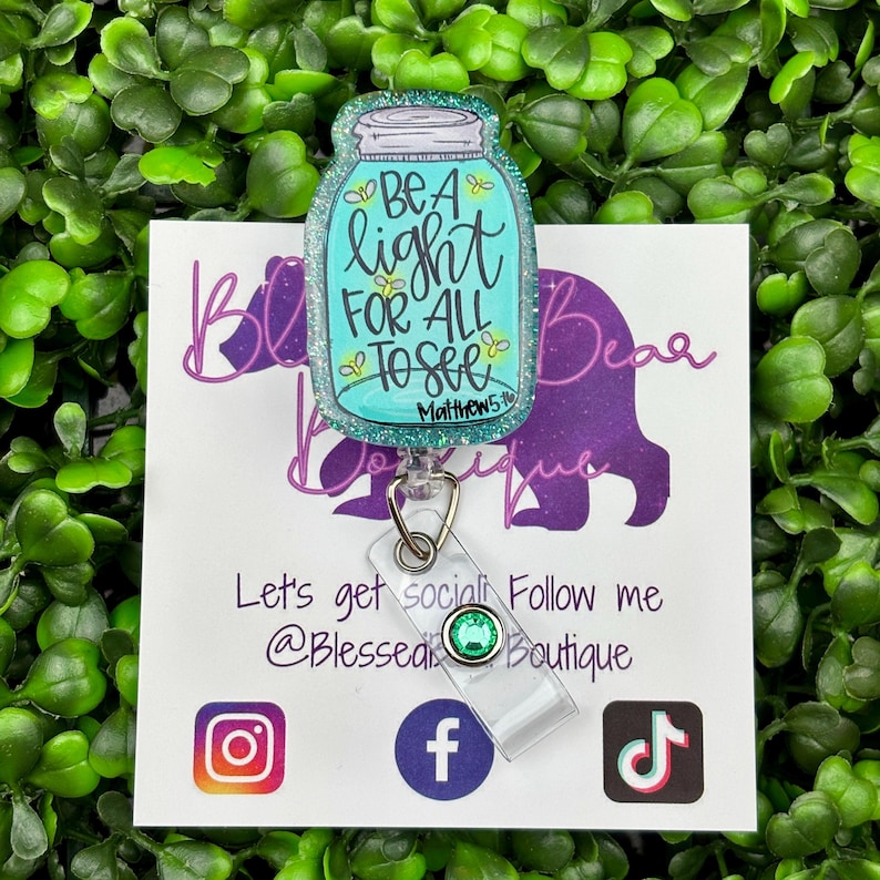The badge reel features a mason jar charm with a turquoise blue glitter background. Inside the jar are the words "BE A light FOR ALL TO SEE" and "Matthew 5:16."