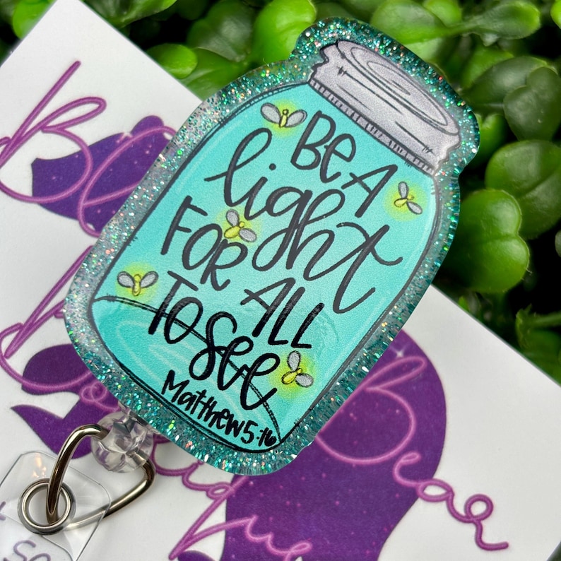 The badge reel features a mason jar charm with a turquoise blue glitter background. Inside the jar are the words "BE A light FOR ALL TO SEE" and "Matthew 5:16."