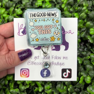 May include: A light blue badge reel with the text "THE GOOD NEWS YOU GOT THIS" and various star and heart illustrations. The reel has a glittery border and a clear plastic clip with a blue gem.