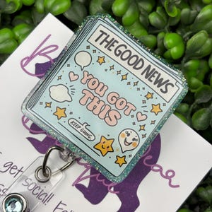 May include: A light blue badge reel with the text "THE GOOD NEWS YOU GOT THIS" and various illustrations. The badge has a glittery border and a clear plastic reel with a metal clip. The words "KEEP GOING" are also visible.