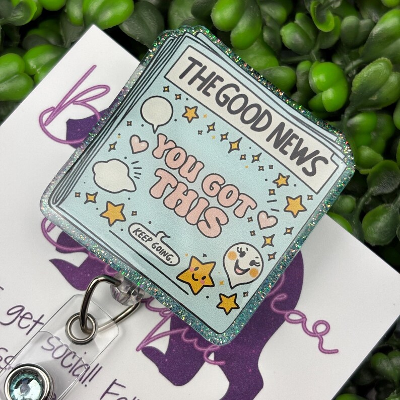 May include: A light blue badge reel with the text "THE GOOD NEWS YOU GOT THIS" and various illustrations. The badge has a glittery border and a clear plastic reel with a metal clip. The words "KEEP GOING" are also visible.