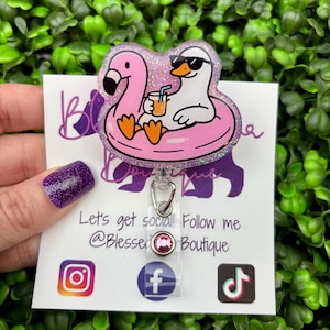 May include: A glittery acrylic badge reel featuring a white duck wearing black sunglasses, sitting in a pink flamingo-shaped pool float while holding an orange drink. The badge reel is attached to a clear plastic strap with a small pink rhinestone.
