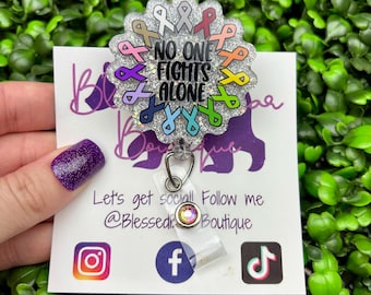 May include: A circular badge reel with a silver glitter finish featuring the text "NO ONE FIGHTS ALONE" in black lettering, surrounded by a border of various colored awareness ribbons. A clear plastic strap with a small crystal accent is attached.
