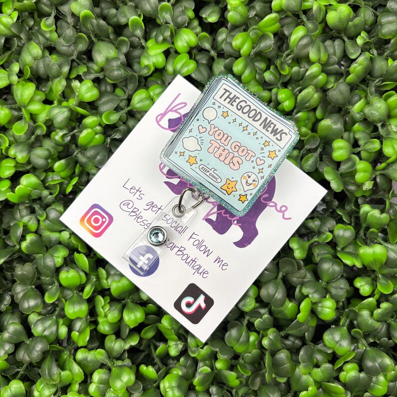 May include: A badge reel with a light blue and glittery design featuring the text "THE GOOD NEWS YOU GOT THIS" and various star and heart illustrations. The reel is attached to a white card with social media icons and text.
