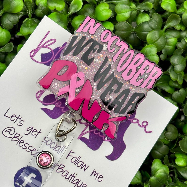 A close-up shot of a hand holding a rectangular retractable badge reel against a background of green artificial leaves. The badge reel has a light pink glitter background. The text "IN OCTOBER WE WEAR PINK" is written in black and bold pink font with a large pink awareness ribbon.