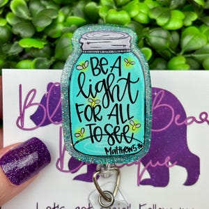 The badge reel features a mason jar charm with a turquoise blue glitter background. Inside the jar are the words "BE A light FOR ALL TO SEE" and "Matthew 5:16."