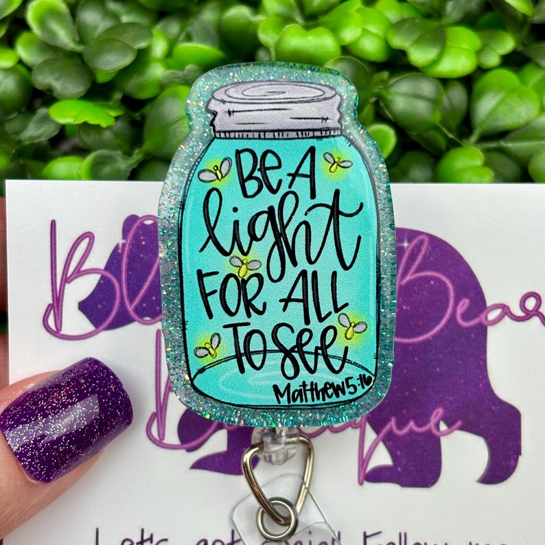 The badge reel features a mason jar charm with a turquoise blue glitter background. Inside the jar are the words "BE A light FOR ALL TO SEE" and "Matthew 5:16."