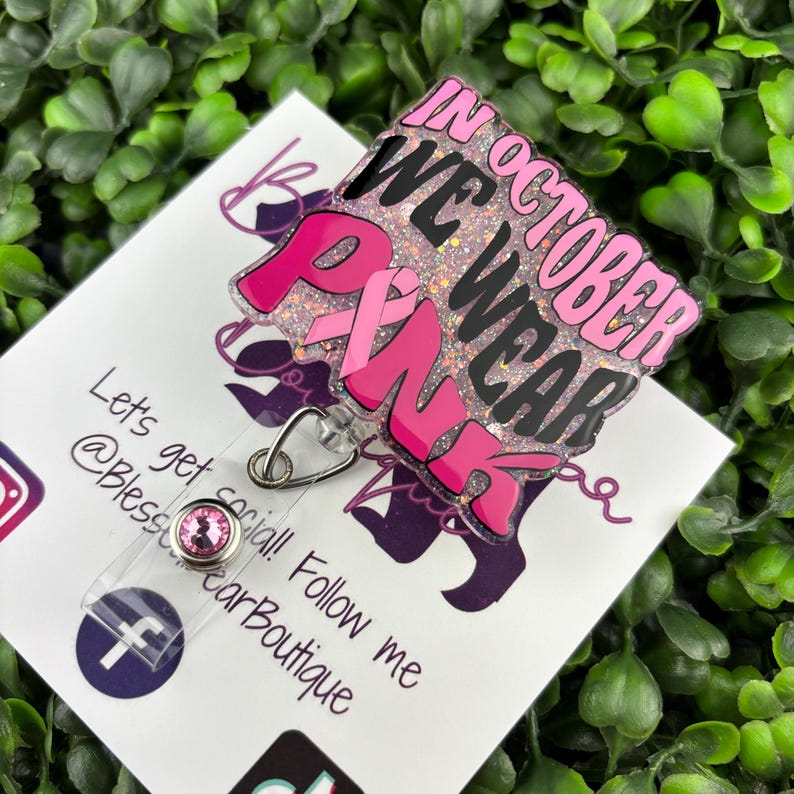 A flat lay image displays a badge reel with the words "IN OCTOBER WE WEAR PINK" in bold, black and pink lettering. A pink ribbon symbol is incorporated into the design. The background of the badge reel is a light pink glitter.