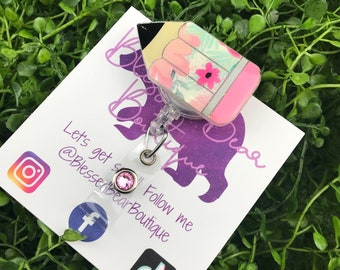 May include: A pink and yellow pencil-shaped retractable badge holder with a floral pattern. The badge holder is attached to a clear plastic retractable cord with a pink rhinestone. The badge holder is on a white card with the text "Let's get social" and social media icons.