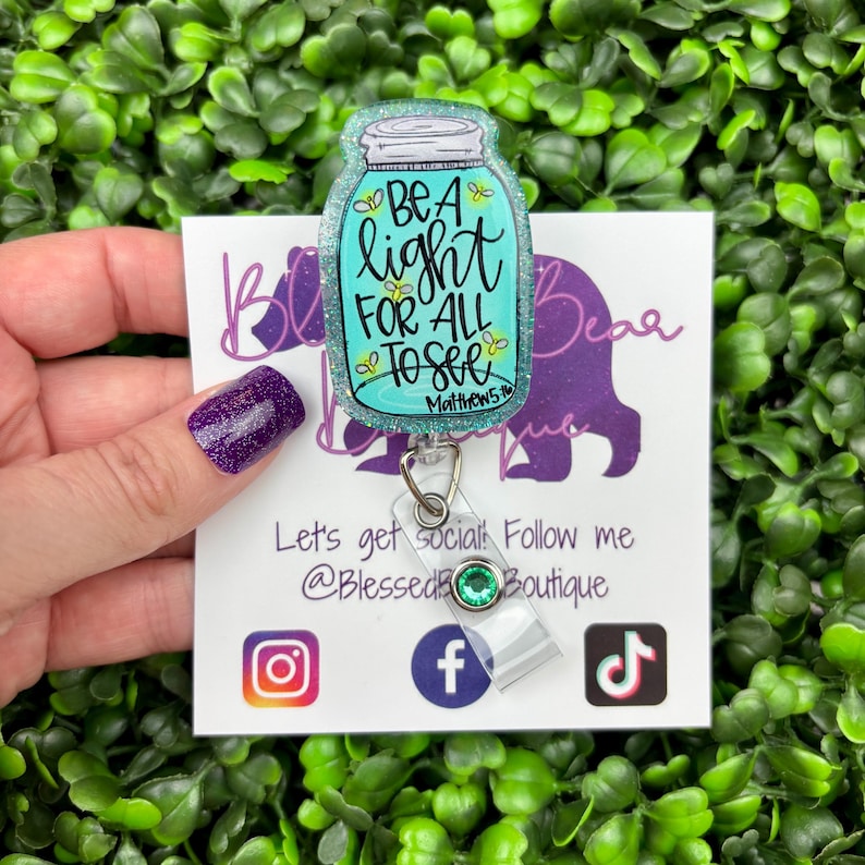 The badge reel features a mason jar charm with a turquoise blue glitter background. Inside the jar are the words "BE A light FOR ALL TO SEE" and "Matthew 5:16."