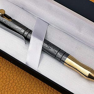 May include: A luxury pen with a patterned black and silver barrel and gold-colored accents. The pen is presented in a black box with a white satin ribbon. The pen's tip and the top button are gold-colored.