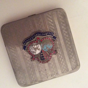May include: A silver cigarette case with a decorative design and a metal emblem featuring the text "Village Portus Douvor" in blue and red.
