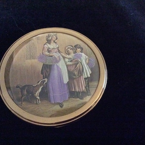 Op de afbeelding: Een rond, goudkleurig compact met een geschilderd beeld van drie mensen en een hond. De mensen zijn gekleed in 18e-eeuwse kleding en houden boeketten lavendel vast. De hond is bruin en zwart en zit op de voorgrond.
