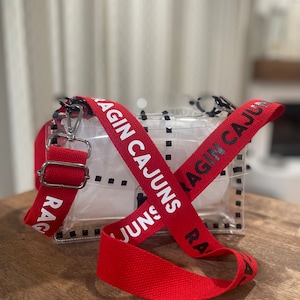 Op de afbeelding: Een transparante crossbody tas van plastic met een rode verstelbare band. De band heeft de woorden "Ragin' Cajuns" in wit erop gedrukt.