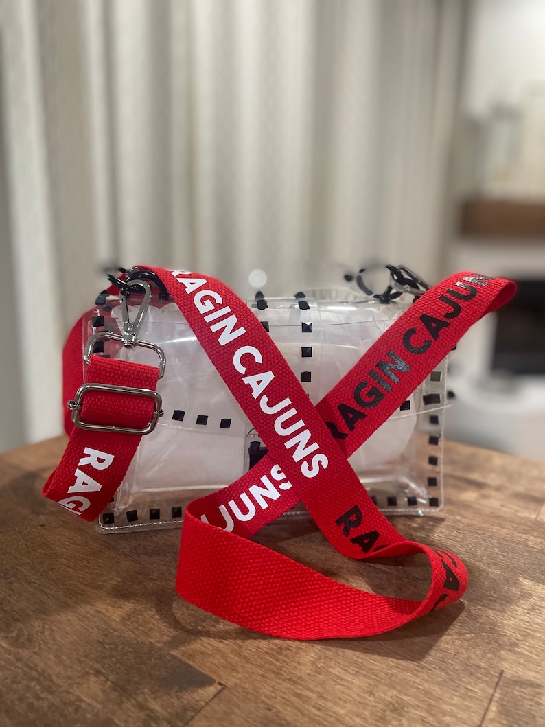 Op de afbeelding: Een transparante crossbody tas van plastic met een rode verstelbare band. De band heeft de woorden "Ragin' Cajuns" in wit erop gedrukt.