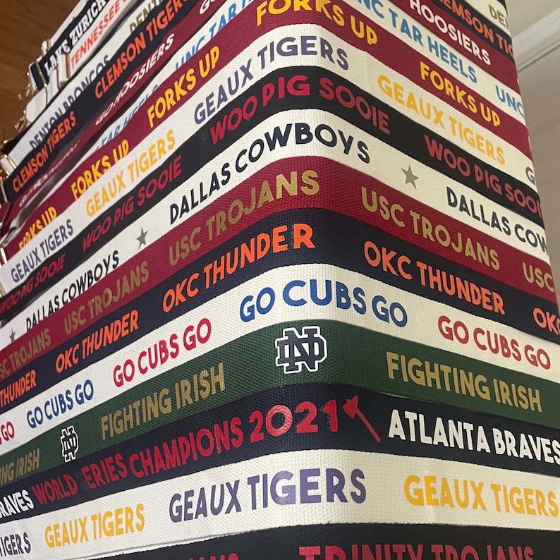 Op de afbeelding: Een close-up van een verzameling kleurrijke stoffen banden met verschillende college- en sportteamlogo's en namen, waaronder "Geaux Tigers", "USC Trojans", "Dallas Cowboys", "OKC Thunder", "Go Cubs Go", "Fighting Irish", "World Series Champions 2021" en "Atlanta Braves".