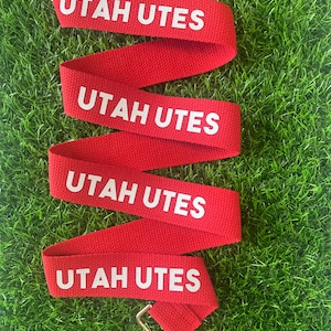 Op de afbeelding: Rode en witte lanyard met de tekst "UTAH UTES" die vier keer wordt herhaald. De lanyard heeft een zilveren metalen clip.