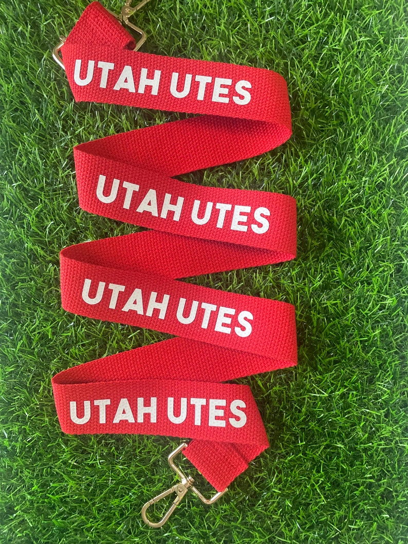 Op de afbeelding: Rode en witte lanyard met de tekst "UTAH UTES" die vier keer wordt herhaald. De lanyard heeft een zilveren metalen clip.