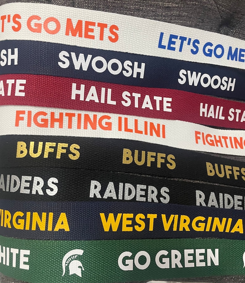 Op de afbeelding: Een close-up van een stapel kleurrijke stoffen polsbandjes met verschillende college-teamlogo's en slogans, waaronder "Let's Go Mets", "Swoosh", "Hail State", "Fighting Illini", "Buffs", "Raiders", "West Virginia" en "Go Green".