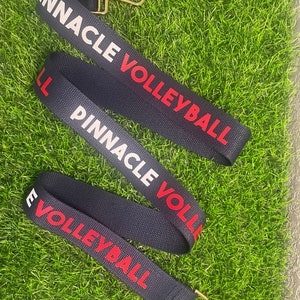 Op de afbeelding: Drie zwart-wit gestreepte banden met gouden gespen. De banden hebben de tekst "Pinnacle Volleyball" in rood en wit gedrukt.