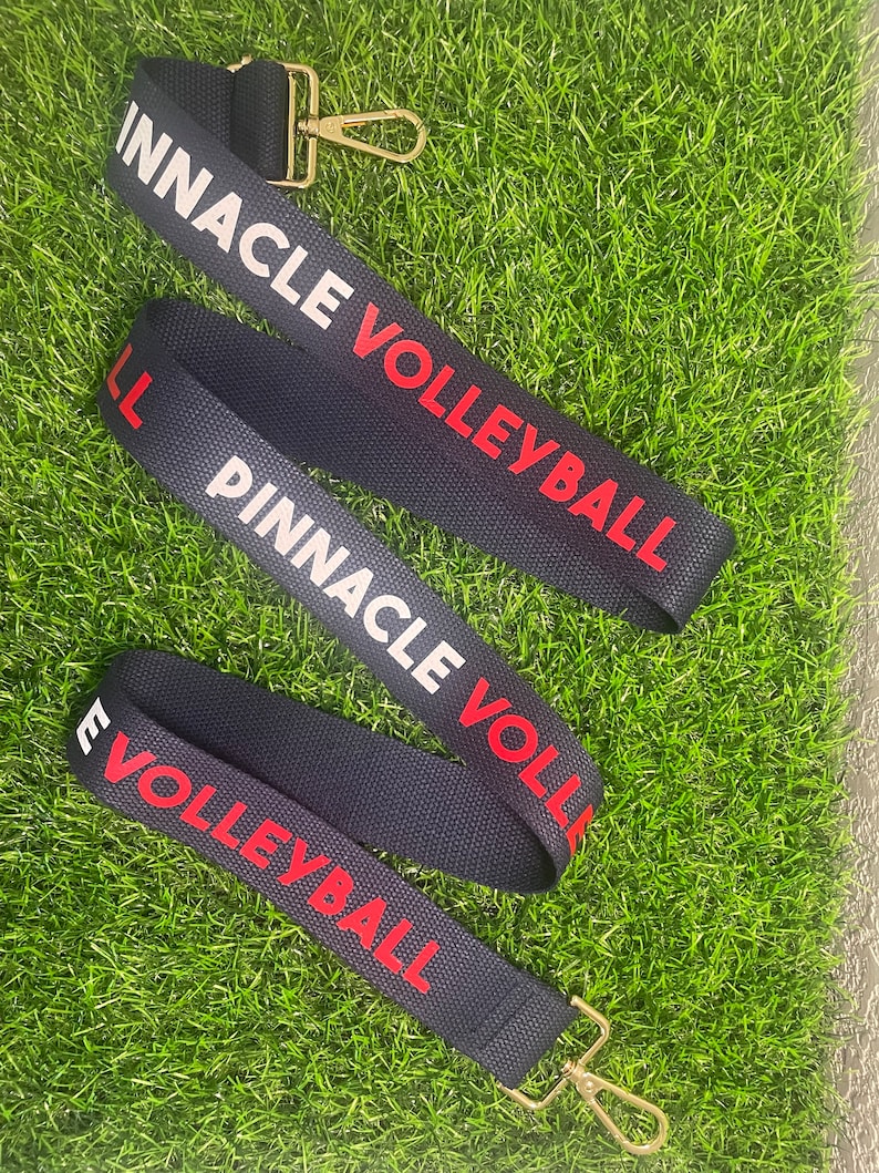Op de afbeelding: Drie zwart-wit gestreepte banden met gouden gespen. De banden hebben de tekst "Pinnacle Volleyball" in rood en wit gedrukt.