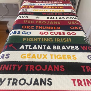 Op de afbeelding: Een close-up van een stapel kleurrijke stoffen polsbandjes met verschillende college- en sportteamlogo's en -namen, waaronder "USC Trojans", "Geaux Tigers", "Fighting Irish", "Go Cubs Go", "OKC Thunder" en "Atlanta Braves".