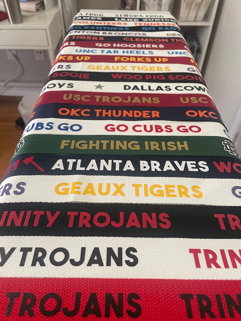 Op de afbeelding: Een close-up van een stapel kleurrijke stoffen polsbandjes met verschillende college- en sportteamlogo's en -namen, waaronder "USC Trojans", "Geaux Tigers", "Fighting Irish", "Go Cubs Go", "OKC Thunder" en "Atlanta Braves".