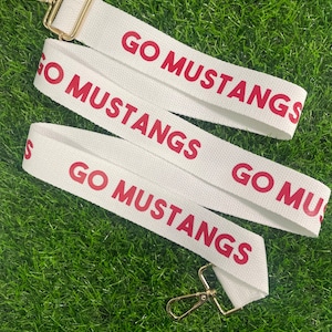 Op de afbeelding: Witte canvasband met gouden metalen gespen. De band heeft de tekst "GO MUSTANGS" in rood gedrukt.