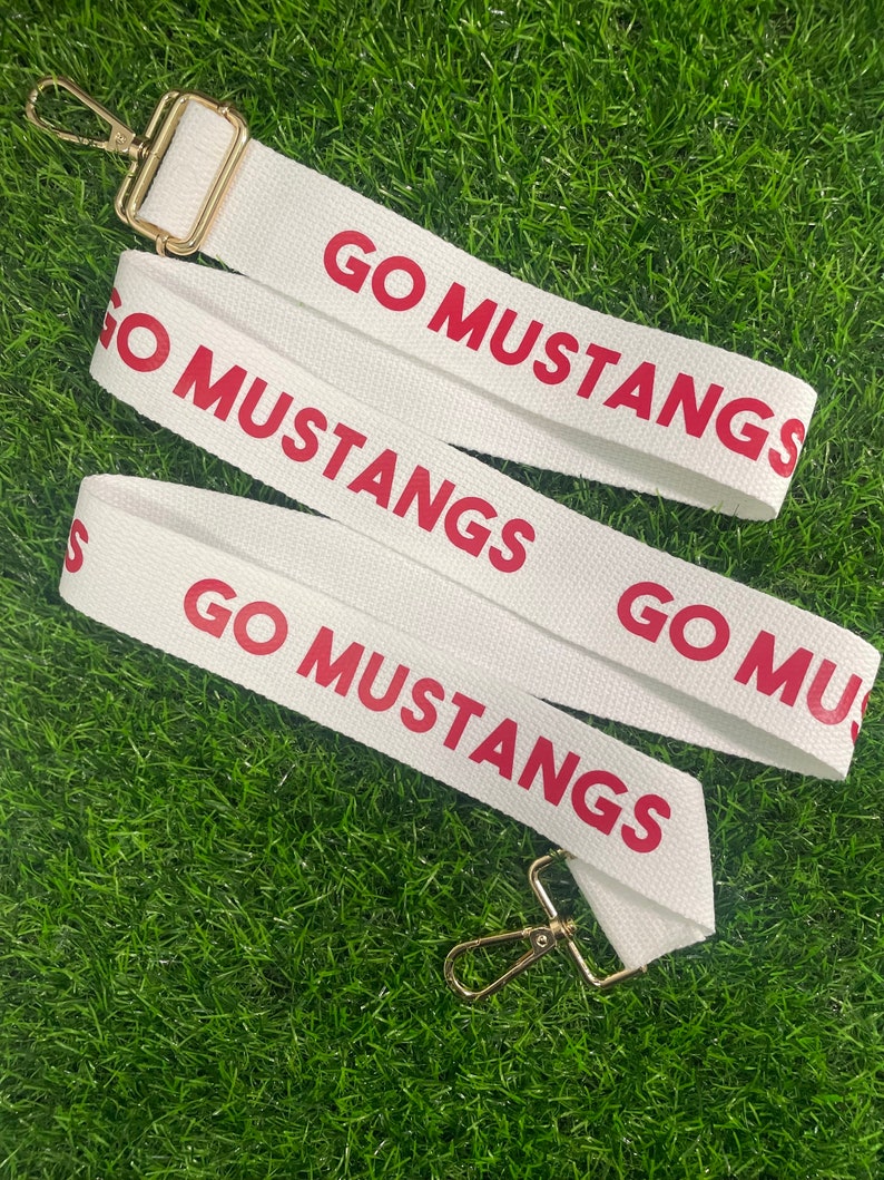 Op de afbeelding: Witte canvasband met gouden metalen gespen. De band heeft de tekst "GO MUSTANGS" in rood gedrukt.
