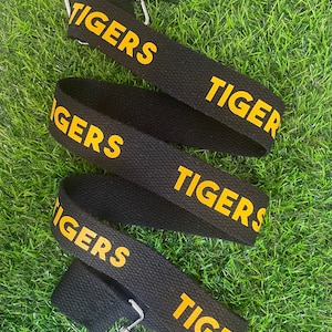 Op de afbeelding: Zwarte verstelbare band met gouden belettering die "TIGERS" herhaalt op de band.