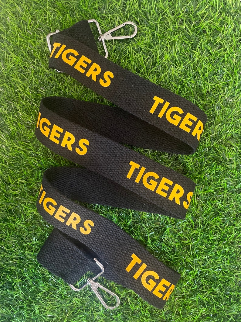 Op de afbeelding: Zwarte verstelbare band met gouden belettering die "TIGERS" herhaalt op de band.