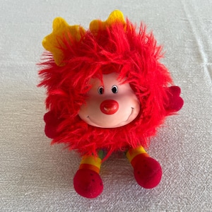 May include: A plush toy with a smiling face, red fur, and yellow star-shaped accents. The toy has red hands and feet, and striped legs in rainbow colors. The toy is designed to look like a friendly creature.