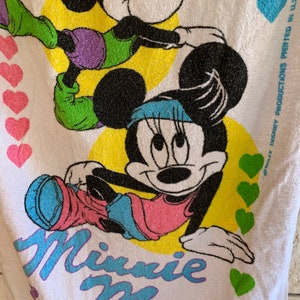 Puede incluir: Una toalla de playa blanca con una imagen de dibujos animados de Minnie Mouse haciendo ejercicio. La toalla tiene un fondo amarillo con corazones verdes y el texto "Minnie Mouse" en letras azules y moradas. El texto "#Walt Disney Productions Printed in U.S.A." est&aacute; impreso en negro en la esquina inferior derecha de la toalla.