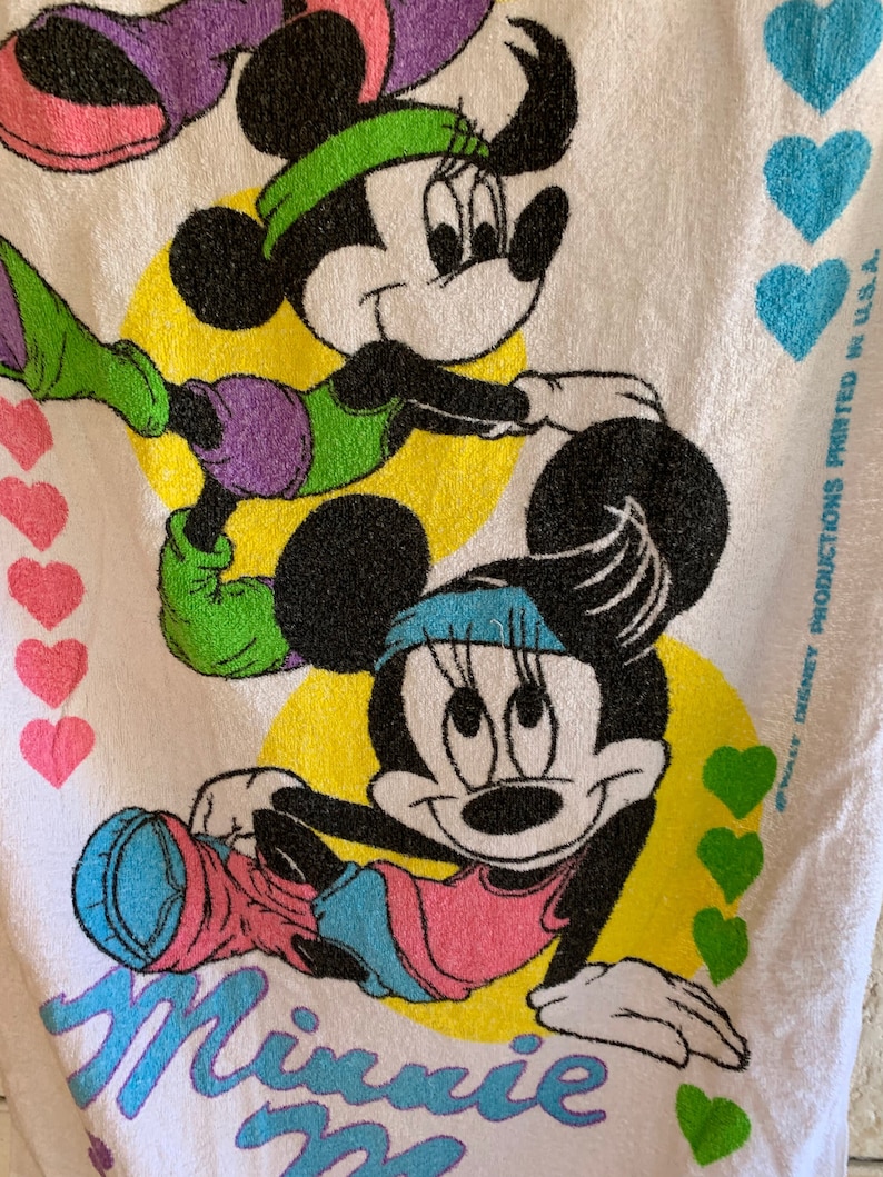Puede incluir: Una toalla de playa blanca con una imagen de dibujos animados de Minnie Mouse haciendo ejercicio. La toalla tiene un fondo amarillo con corazones verdes y el texto "Minnie Mouse" en letras azules y moradas. El texto "#Walt Disney Productions Printed in U.S.A." est&aacute; impreso en negro en la esquina inferior derecha de la toalla.