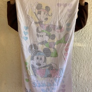 Puede incluir: Una toalla de ba&ntilde;o blanca con una imagen de dibujos animados rosa, verde y azul de Mickey Mouse y Minnie Mouse. La toalla tiene el texto "Mouse" y "Minnie" impreso en ella. La toalla est&aacute; decorada con corazones rosas.