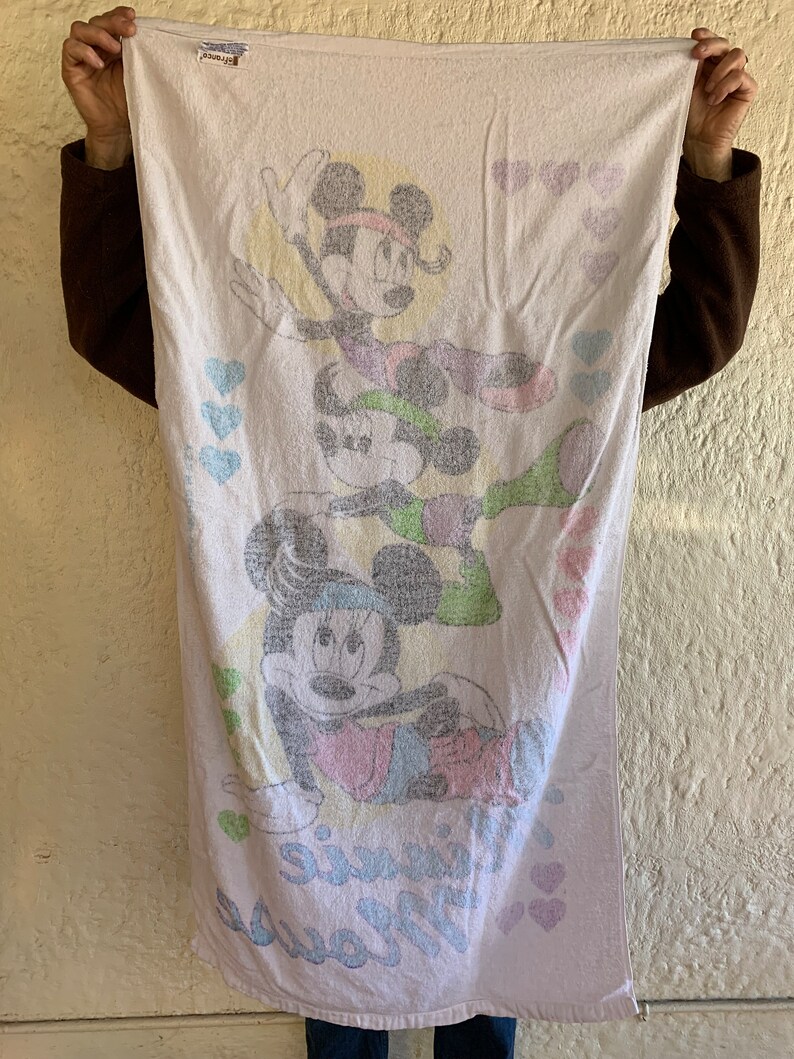 Puede incluir: Una toalla de ba&ntilde;o blanca con una imagen de dibujos animados rosa, verde y azul de Mickey Mouse y Minnie Mouse. La toalla tiene el texto "Mouse" y "Minnie" impreso en ella. La toalla est&aacute; decorada con corazones rosas.