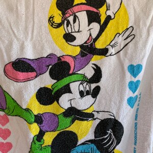 Puede incluir: Una toalla blanca con tres im&aacute;genes de dibujos animados de Minnie Mouse con atuendos de entrenamiento coloridos. La toalla tiene un fondo amarillo con corazones morados alrededor de los bordes. El texto "&copy; WALT DISNEY PRODUCTIONS PRINTED IN U.S.A." est&aacute; impreso en la esquina inferior derecha.