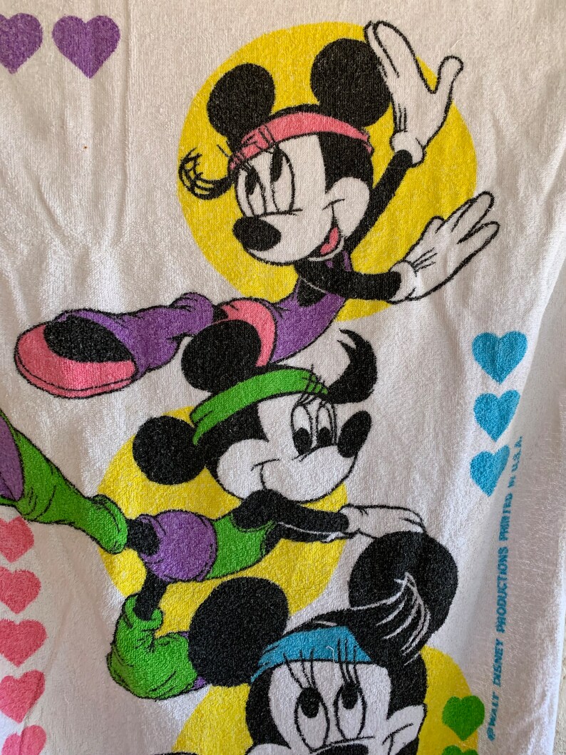 Puede incluir: Una toalla blanca con tres im&aacute;genes de dibujos animados de Minnie Mouse con atuendos de entrenamiento coloridos. La toalla tiene un fondo amarillo con corazones morados alrededor de los bordes. El texto "&copy; WALT DISNEY PRODUCTIONS PRINTED IN U.S.A." est&aacute; impreso en la esquina inferior derecha.